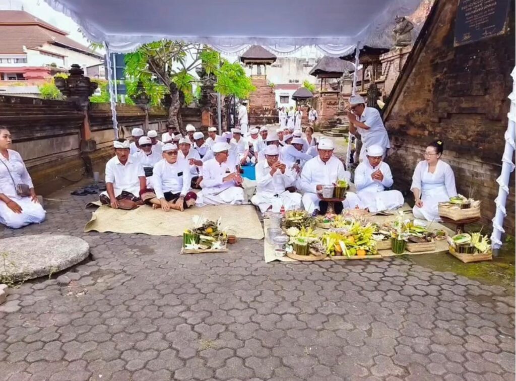 Whatsapp image 20260607 at 21 23 52 catrawarta Group of people in white attire sit on mats under a canopy at a temple preparing offerings with fruit and palm leaf trays